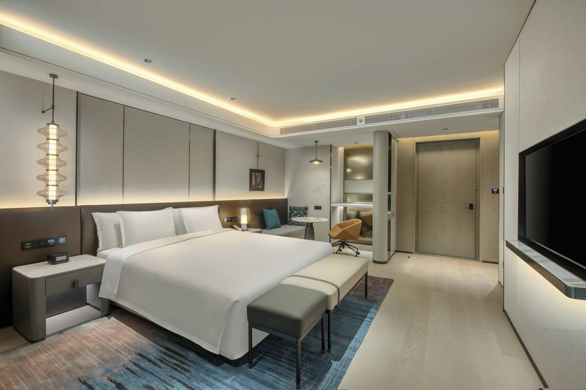 A modern hotel room with a large bed, bedside tables, a bench, a TV, a desk, and a wardrobe.