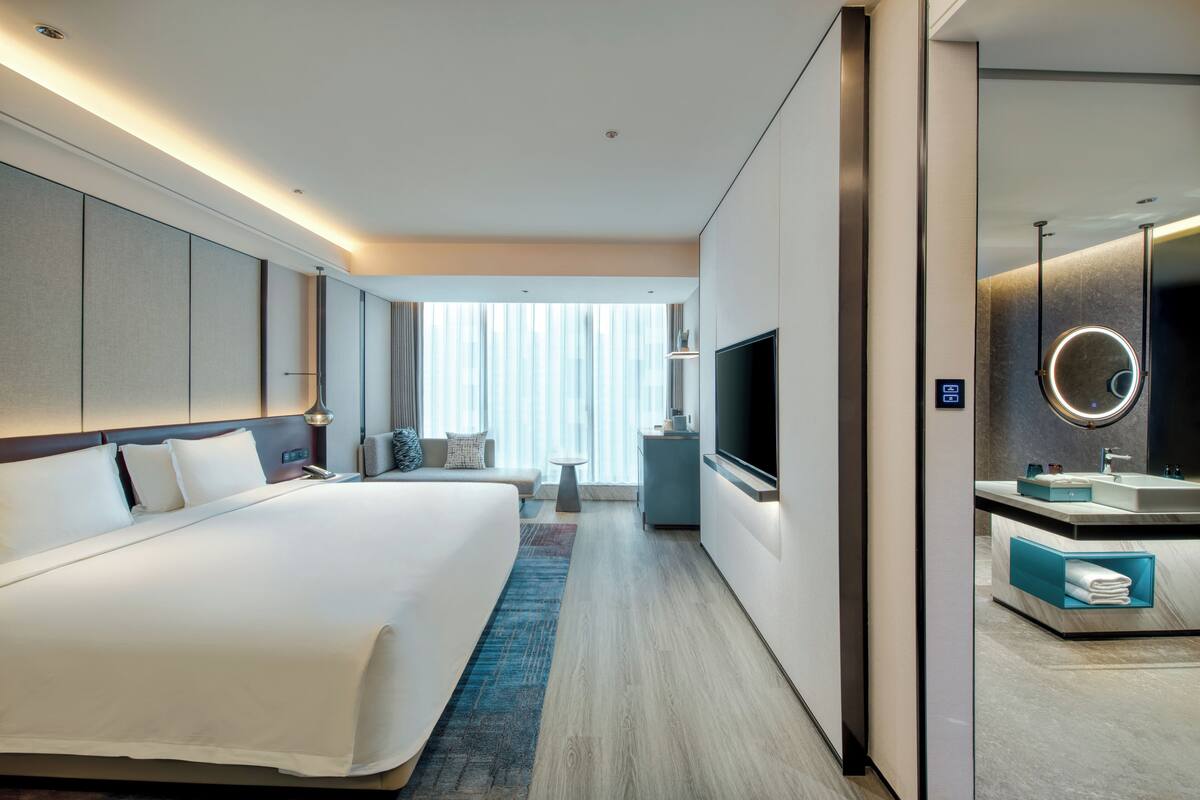 A modern hotel room with a large bed, a flat-screen TV, a vanity with a mirror, and a small seating area with a table and chairs.