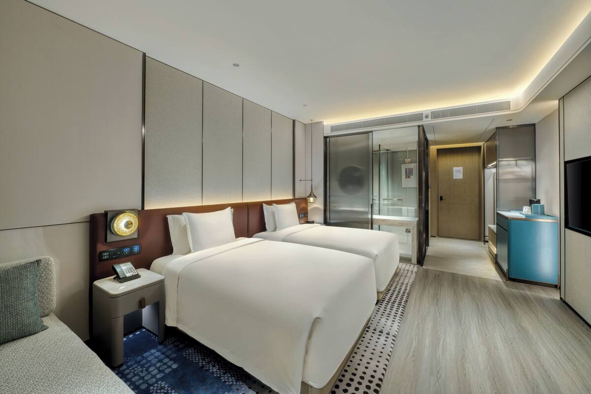 A hotel room with two beds, a desk with a phone, a TV, and a bathroom with a mirror and sink.