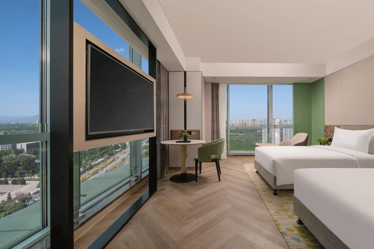 A modern hotel room with a large TV, two beds, a small table, and a view of the city.