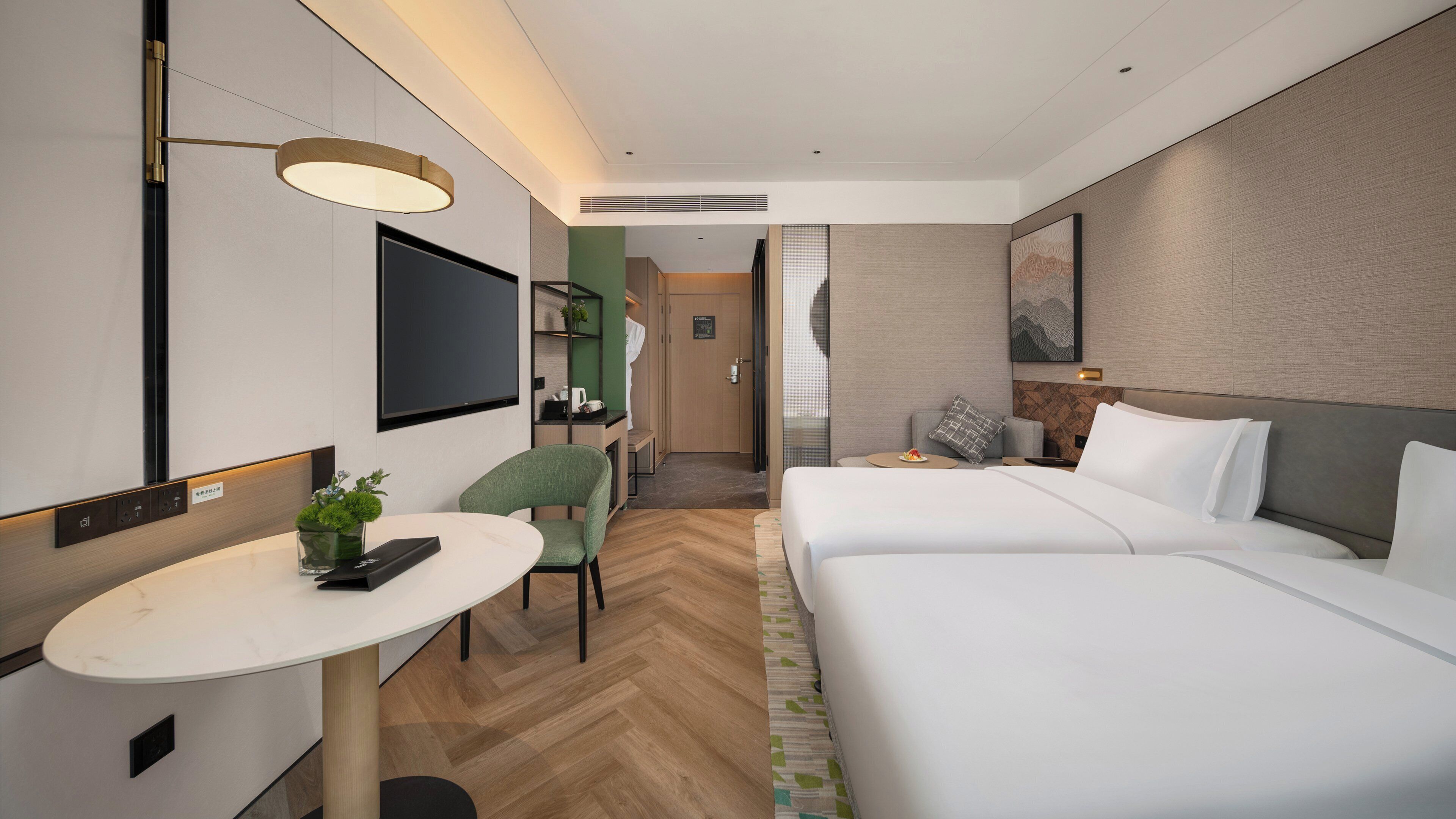 A hotel room with a bed, a desk, a chair, a TV, and a small table with a plant.