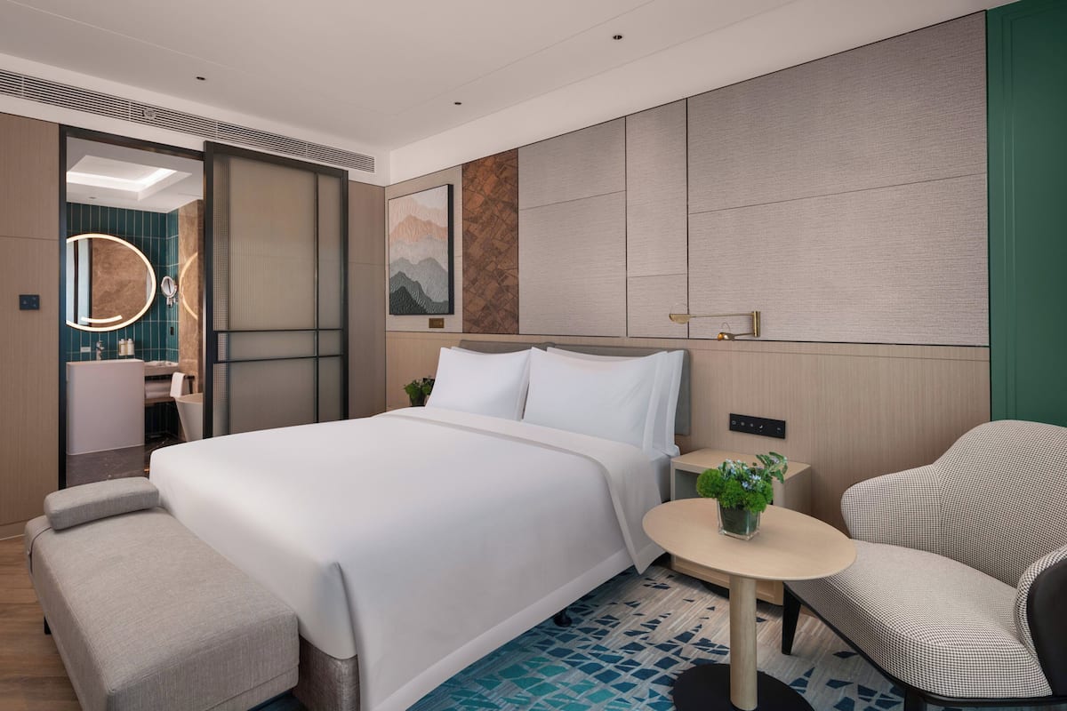 A modern hotel room with a large bed, a small round table, a chair, and a bathroom visible through an open door.