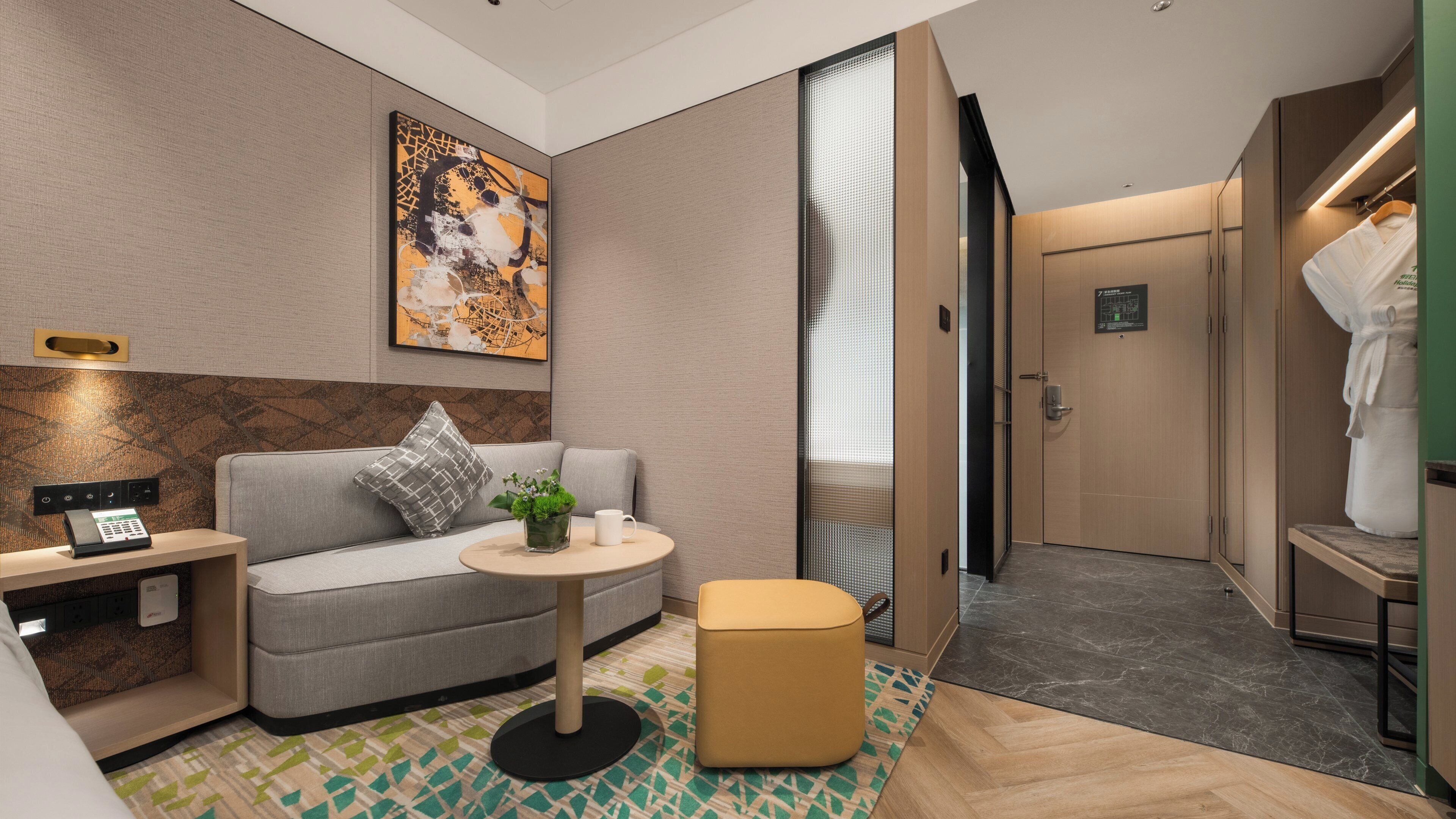 Una habitación de hotel moderna con sofá, una mesita, una planta decorativa y una bata colgada en la puerta.
