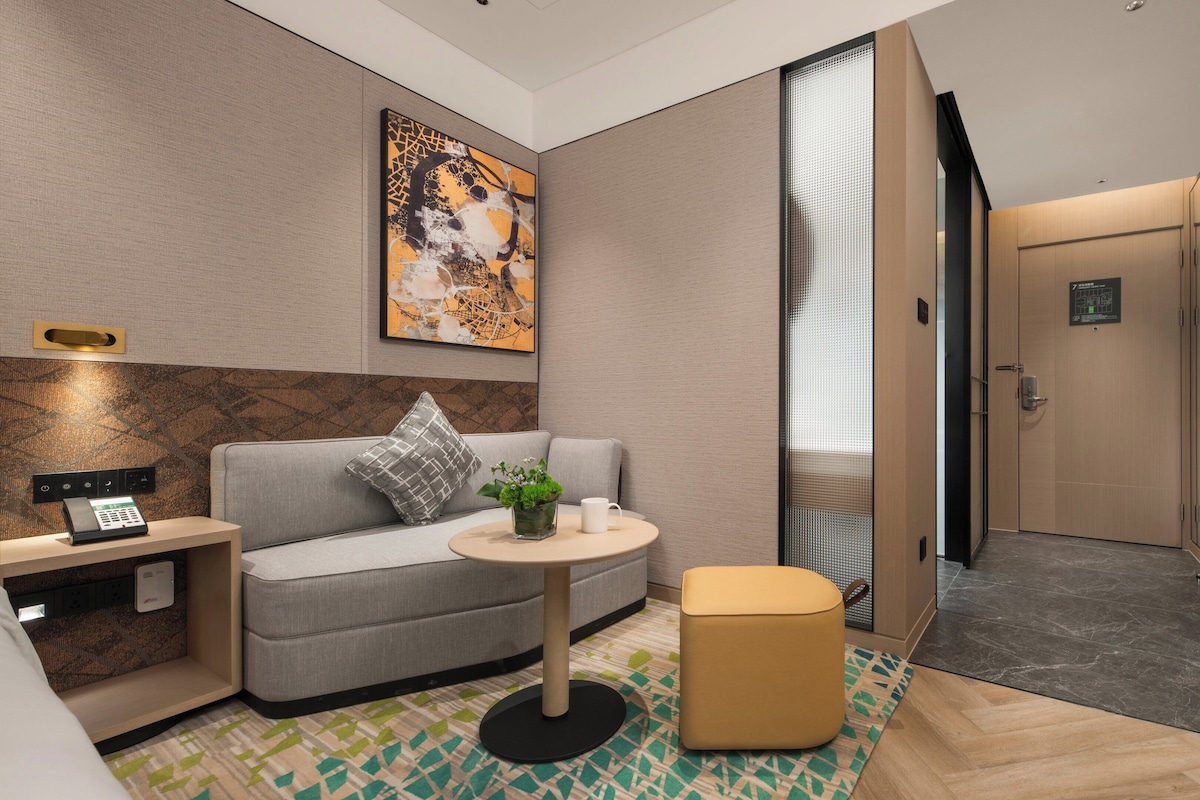 A modern hotel room with a sofa, a small table, a decorative plant, and a robe hanging on the door.
