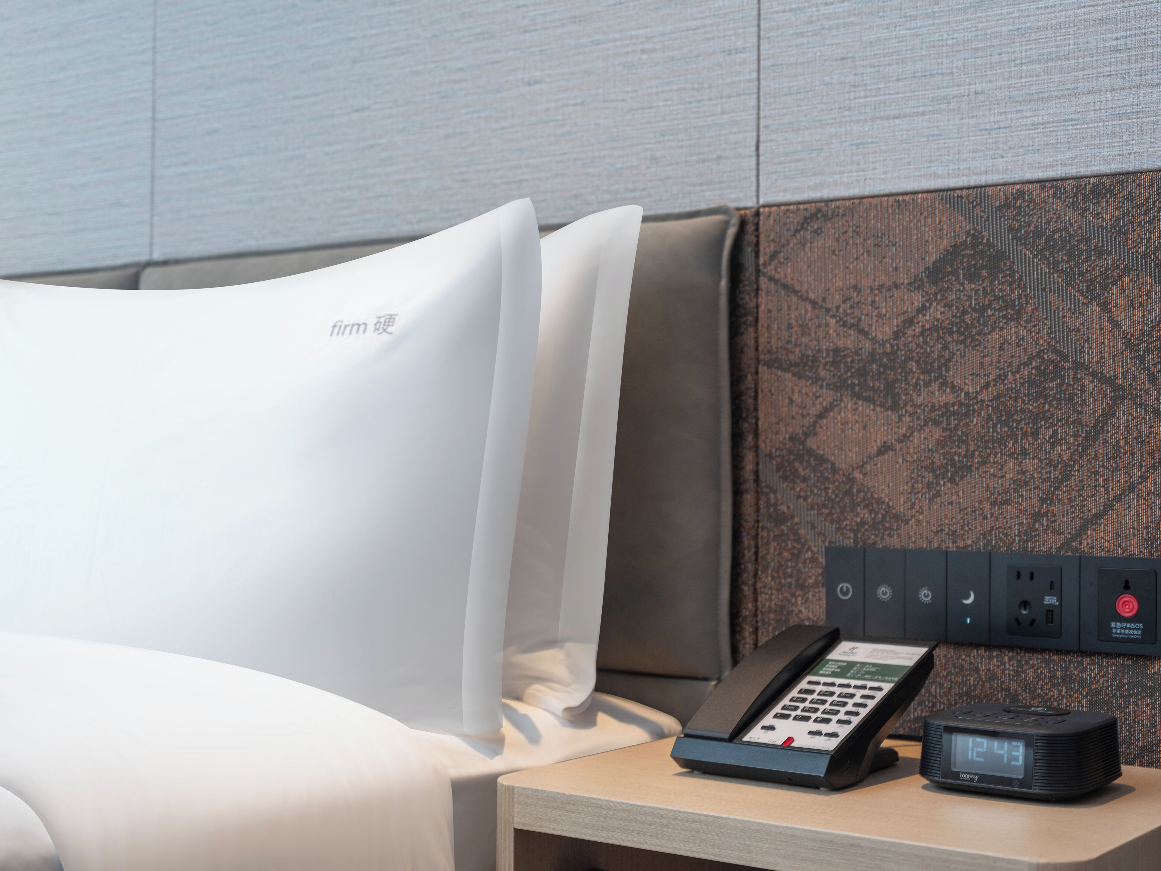 Habitación de hotel con cama, mesita de noche con teléfono y reloj digital, y pared con textura.