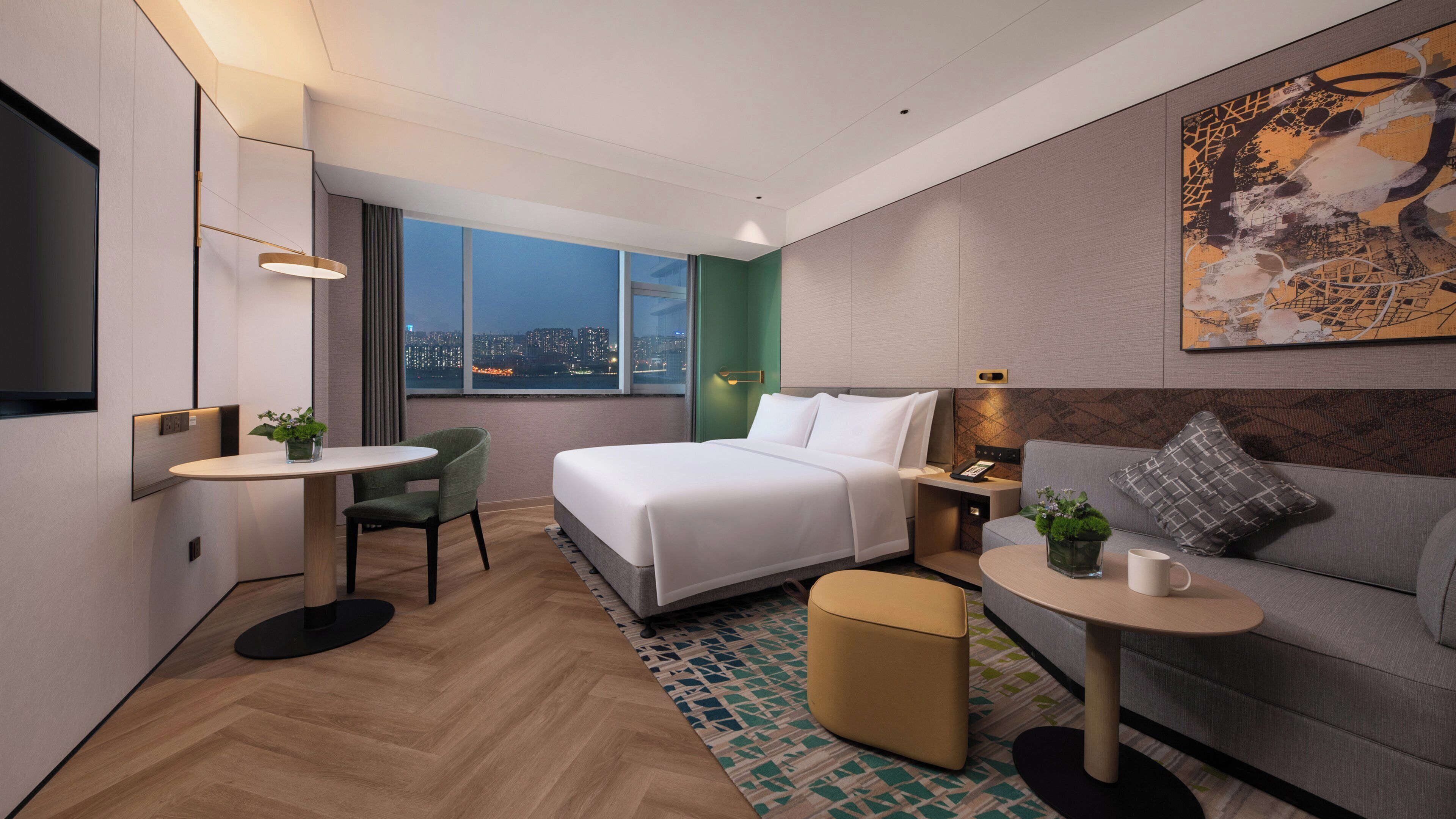 Habitación de hotel moderna con cama, sofá, una mesita, una silla y vista a la ciudad de noche.