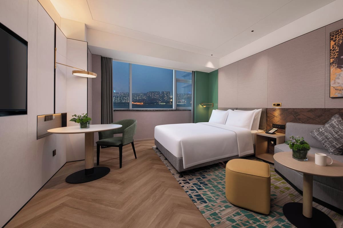 A modern hotel room with a large bed, a sitting area with a sofa and armchair, a small dining table, and a large window offering a city view.