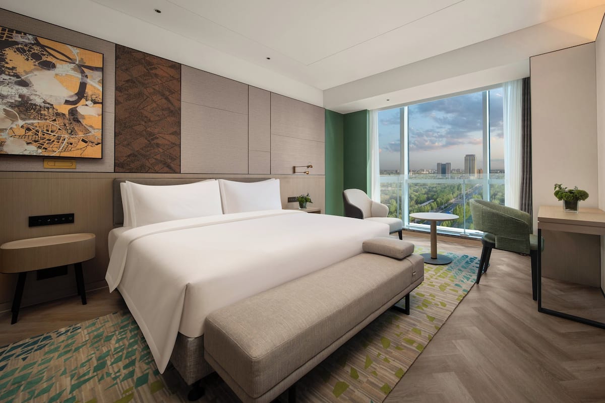 A modern hotel room with a large bed, a desk, a chair, and a view of the city through large windows.