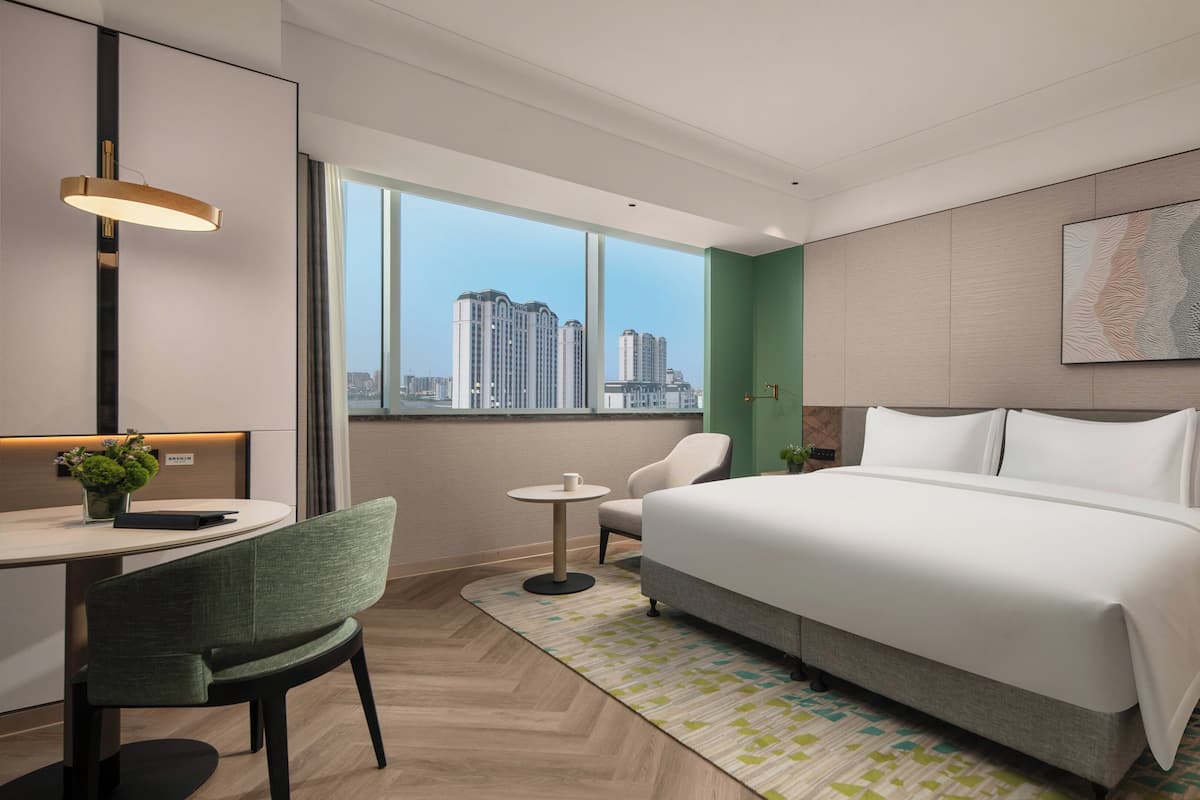 A modern hotel room with a large bed, a desk, a chair, and a view of the cityscape through the window.
