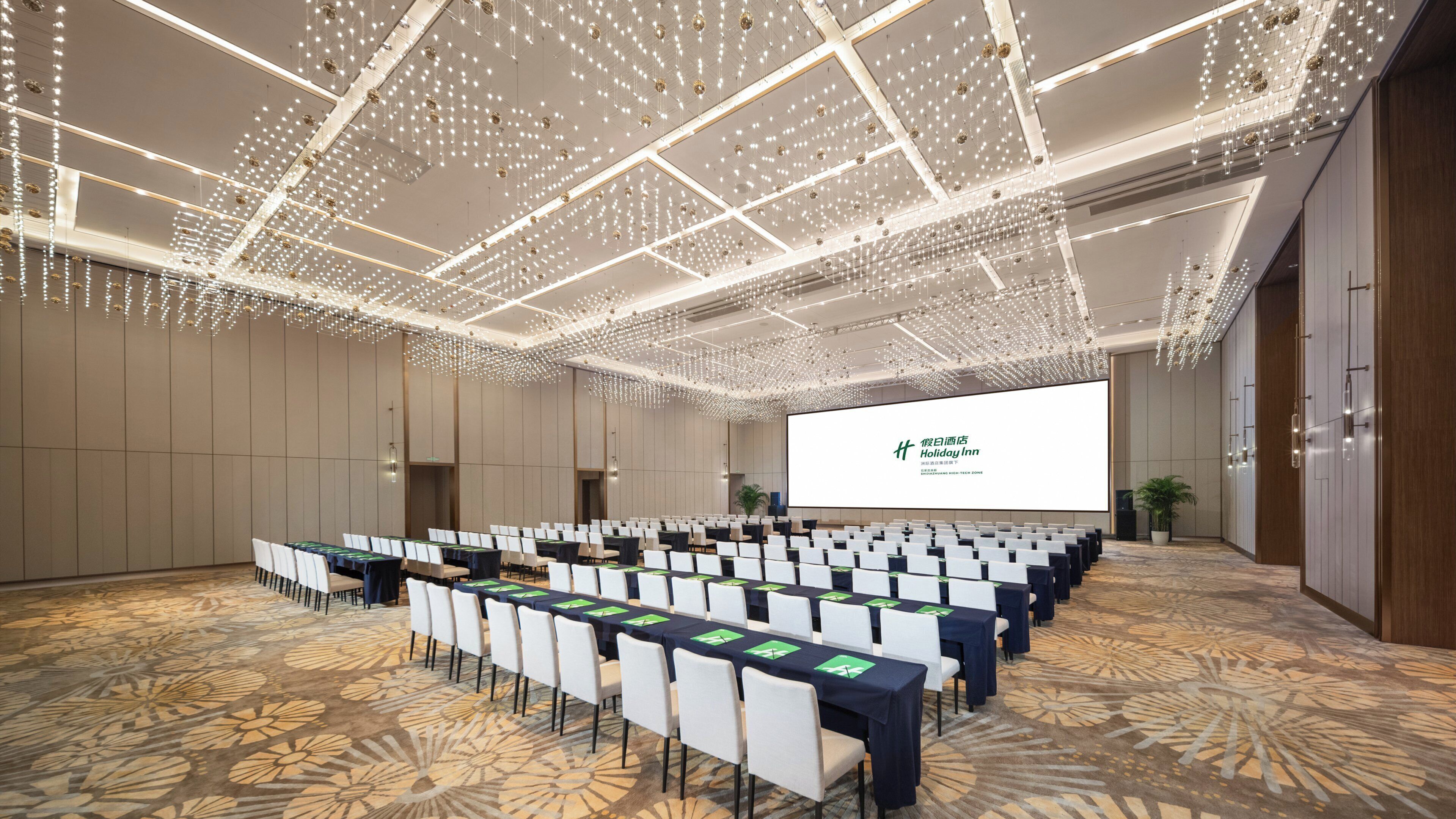 A conference room with rows of tables and chairs, a large screen, and a ceiling with hanging lights.