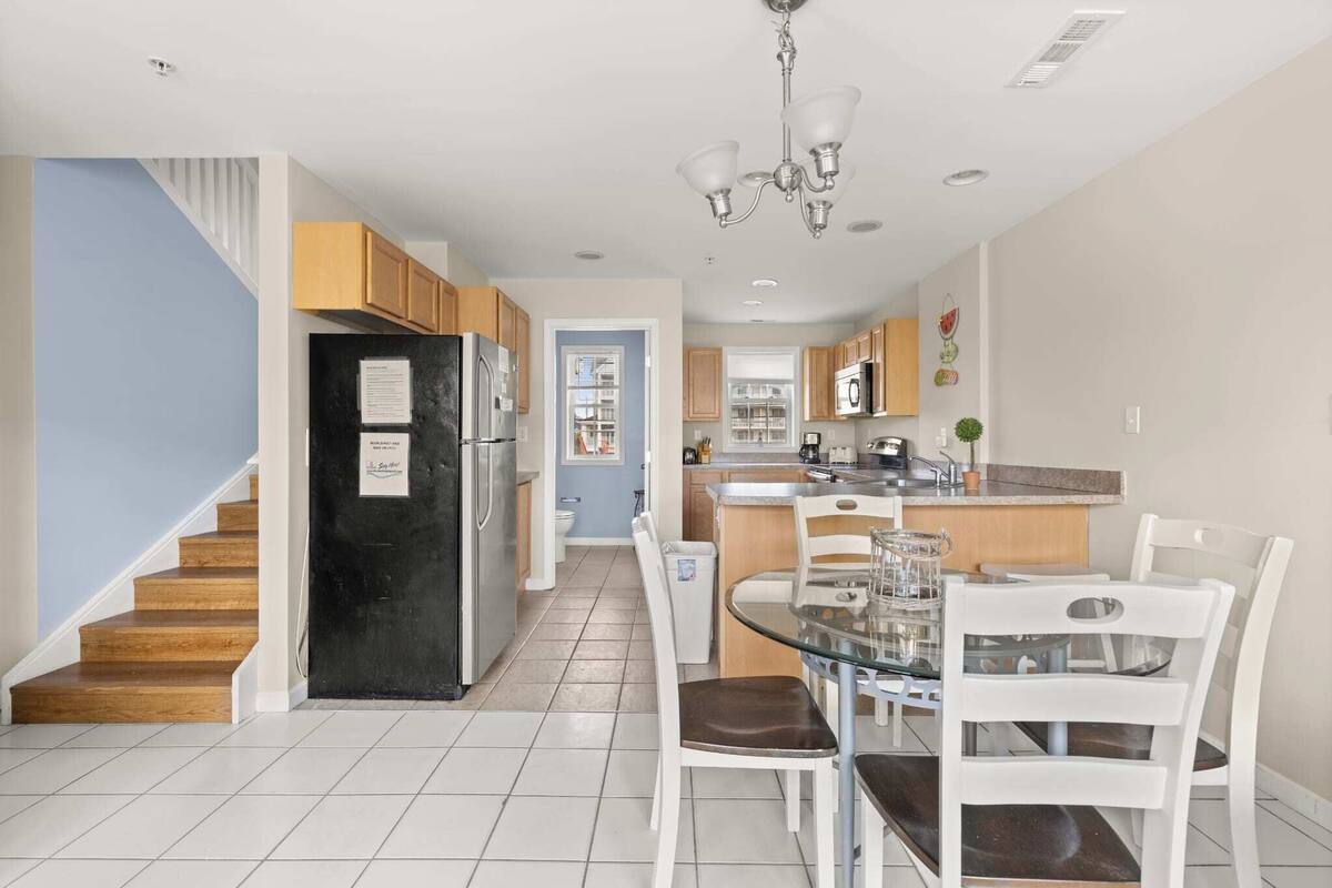 Una cocina moderna con comedor, armarios de madera, un refrigerador negro y una mesa de vidrio con sillas blancas.