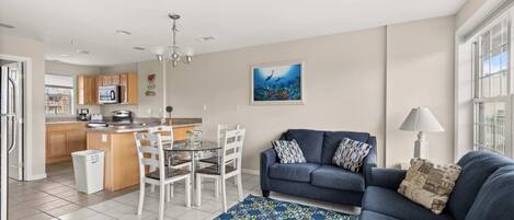 A modern living room with a blue sofa, a dining table with chairs, a kitchen area with wooden cabinets, and a large window.