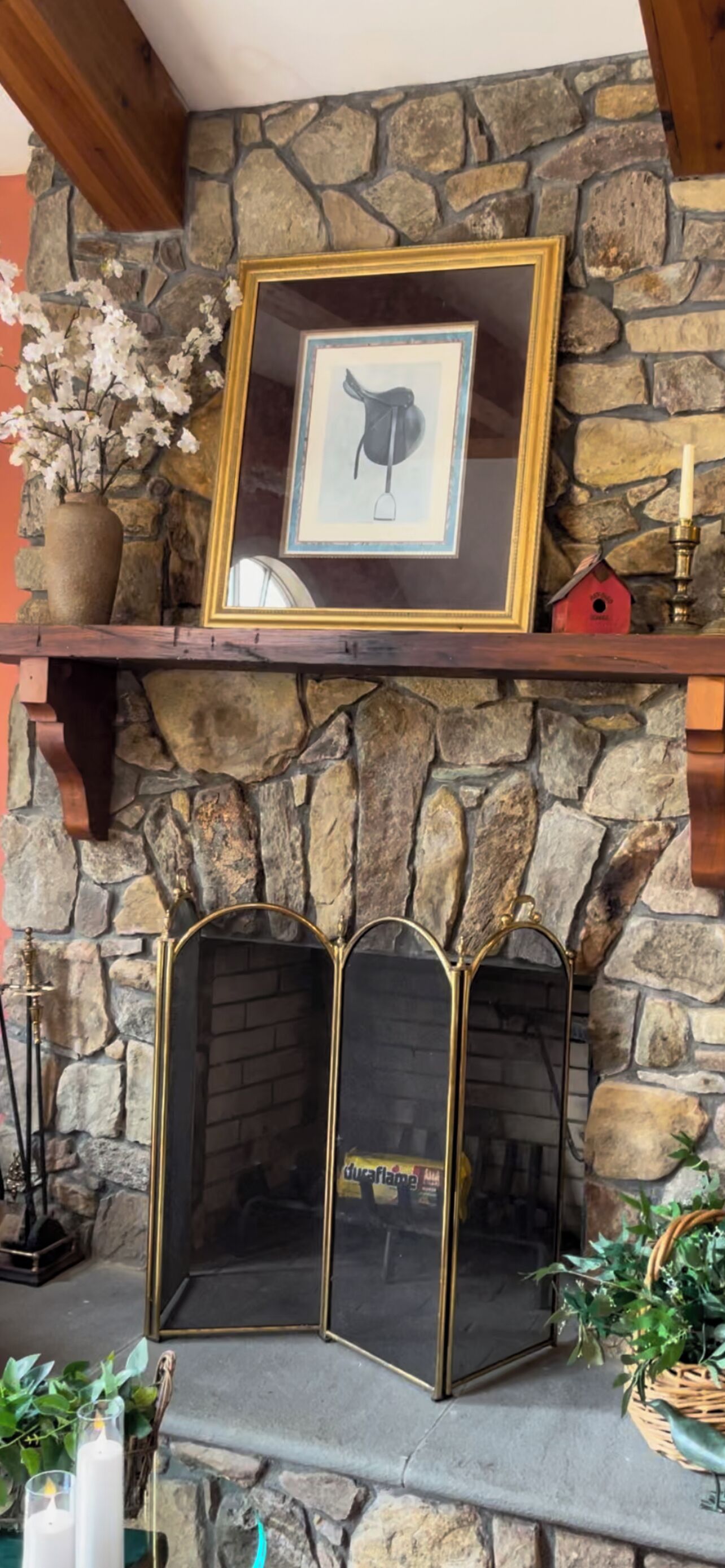 Una chimenea de piedra con un cuadro enmarcado encima, un comedero rojo para pájaros y un candelabro.