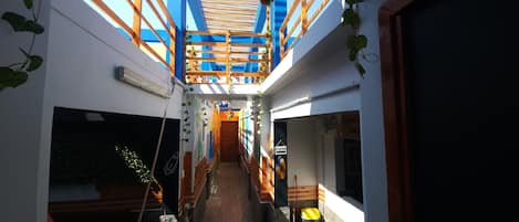 Un estrecho pasillo con vigas de madera y plantas colgantes, que conduce a una puerta con la etiqueta 'SALIDA'.