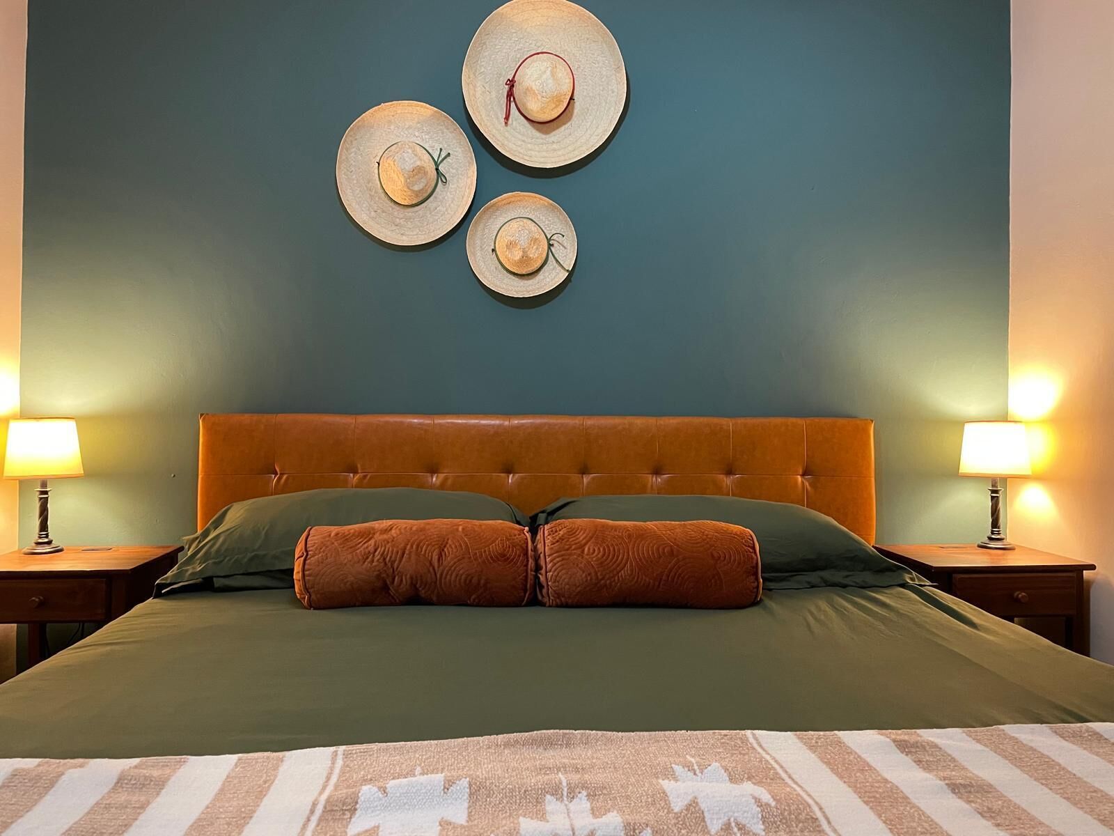 A bedroom with a tufted headboard, a green bedspread, two brown pillows, and a striped area rug. There are two bedside tables with lamps, and three wall-mounted hats.