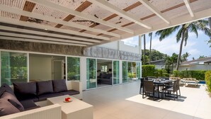 A modern outdoor living area with a sofa, dining table, and chairs, surrounded by glass walls and a view of the outdoors. - Exclusive beach apartment with Wonderful View (Ko Lanta Yai)