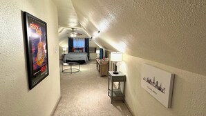 A hotel hallway with a poster of a rock concert, a bed, a desk with a lamp, and a framed cityscape.
