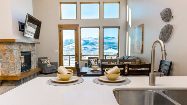A modern kitchen with a stone fireplace, large windows, and a dining area with bowls and plates. - Adventure Awaits at R202 | Ridge Haven in Eden, UT! (Eden)