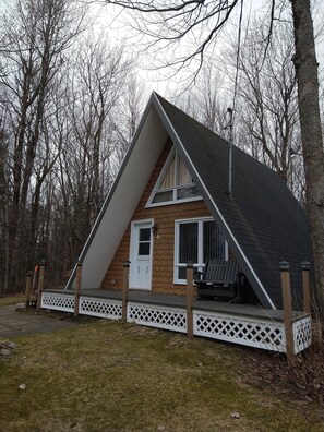 Une petite cabane triangulaire en bois, avec une porte et des fenêtres blanches, entourée d’une clôture en piquets blancs et dotée d’une terrasse.