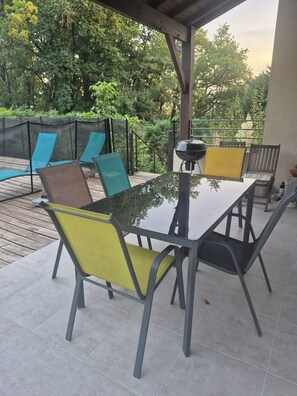 An outdoor patio with a glass table and chairs, a barbecue grill, and a pool in the background.