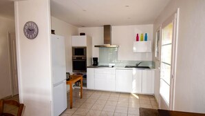 A modern kitchen with white cabinetry, a built-in oven, a microwave, and a coffee maker. There is a wooden table and chairs, a clock on the wall, and a window with a view. - Villa Pech DE Durand Villeneuve SUR LOT Acces PMR (Villeneuve-sur-Lot)