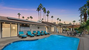 Una piscina con sillones de descanso, palmeras y un patio con mobiliario exterior.