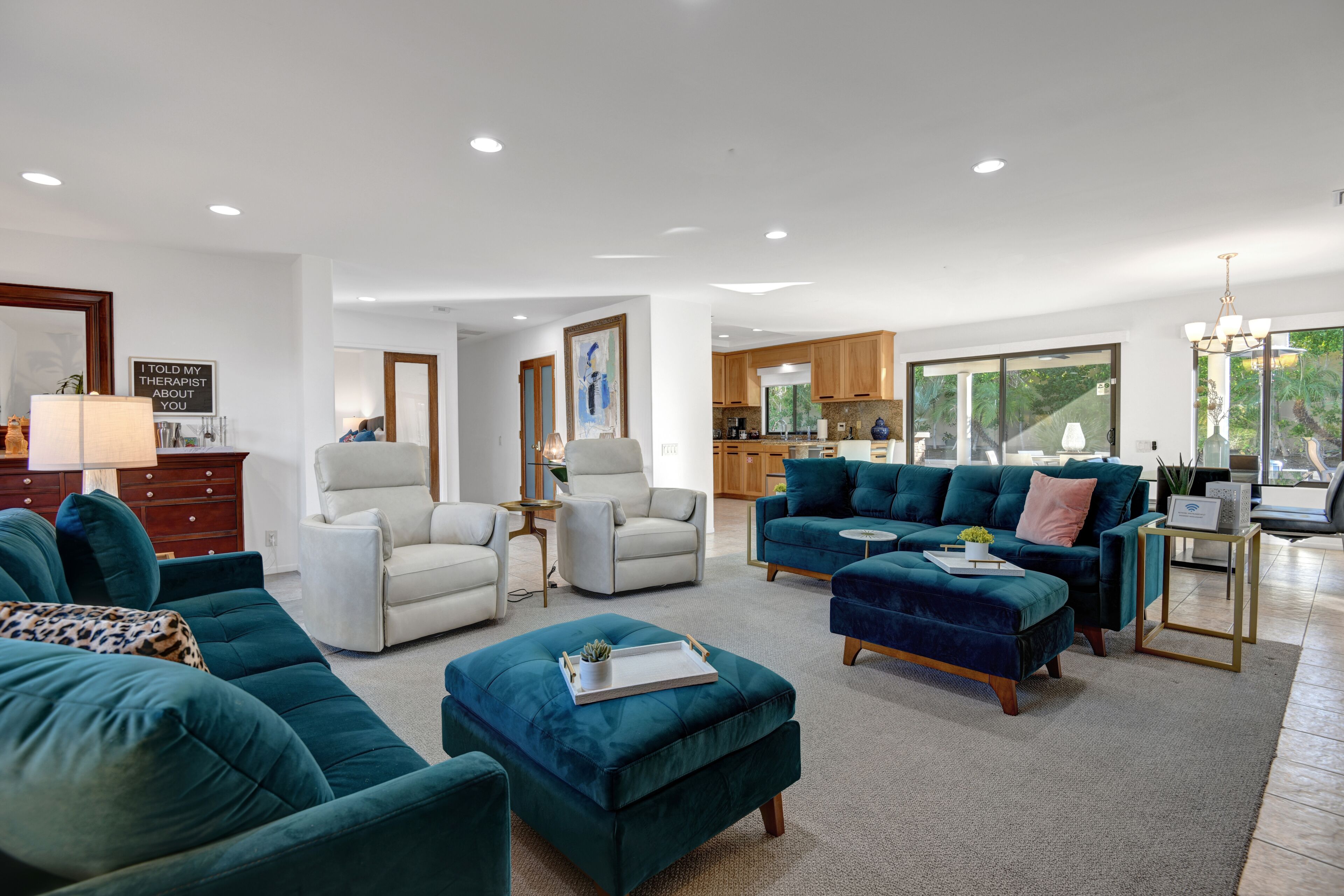 Ein geräumiges Wohnzimmer mit türkisfarbenen Sofas, einem weißen Sessel und einem Couchtisch. Im Hintergrund ist eine Küche mit Holzschränken und großen Fenstern zu sehen.