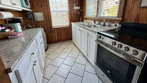 A kitchen with white cabinets, a stainless steel stove, and a window with blinds. - Sea Captain's Cottage 2; Oceanfront Cottage Court (Kill Devil Hills)