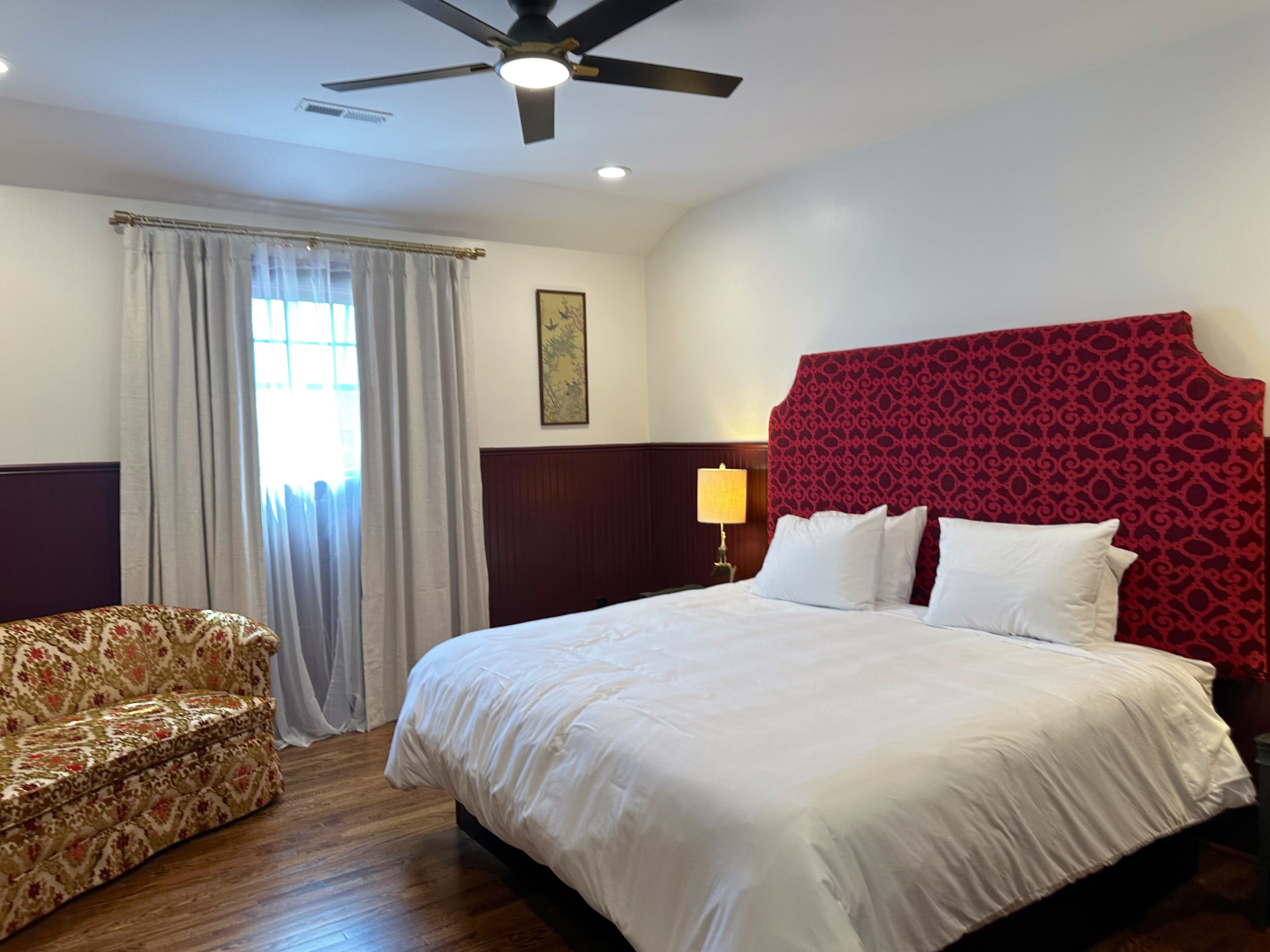 A hotel room with a large bed, a wooden floor, a ceiling fan, a window with curtains, and a patterned armchair.