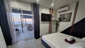 Ein Schlafzimmer mit Bett, Fernseher, einem Fenster mit Vorhängen und Blick ins Freie.