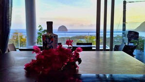Ein Tisch mit einer Vase voller pinkfarbener Blumen und einer Flasche, mit Blick auf eine malerische Meereslandschaft und eine Brücke.