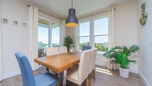 Un coin repas avec une table en bois, des chaises bleues et beiges, un luminaire suspendu et de grandes fenêtres avec des rideaux.