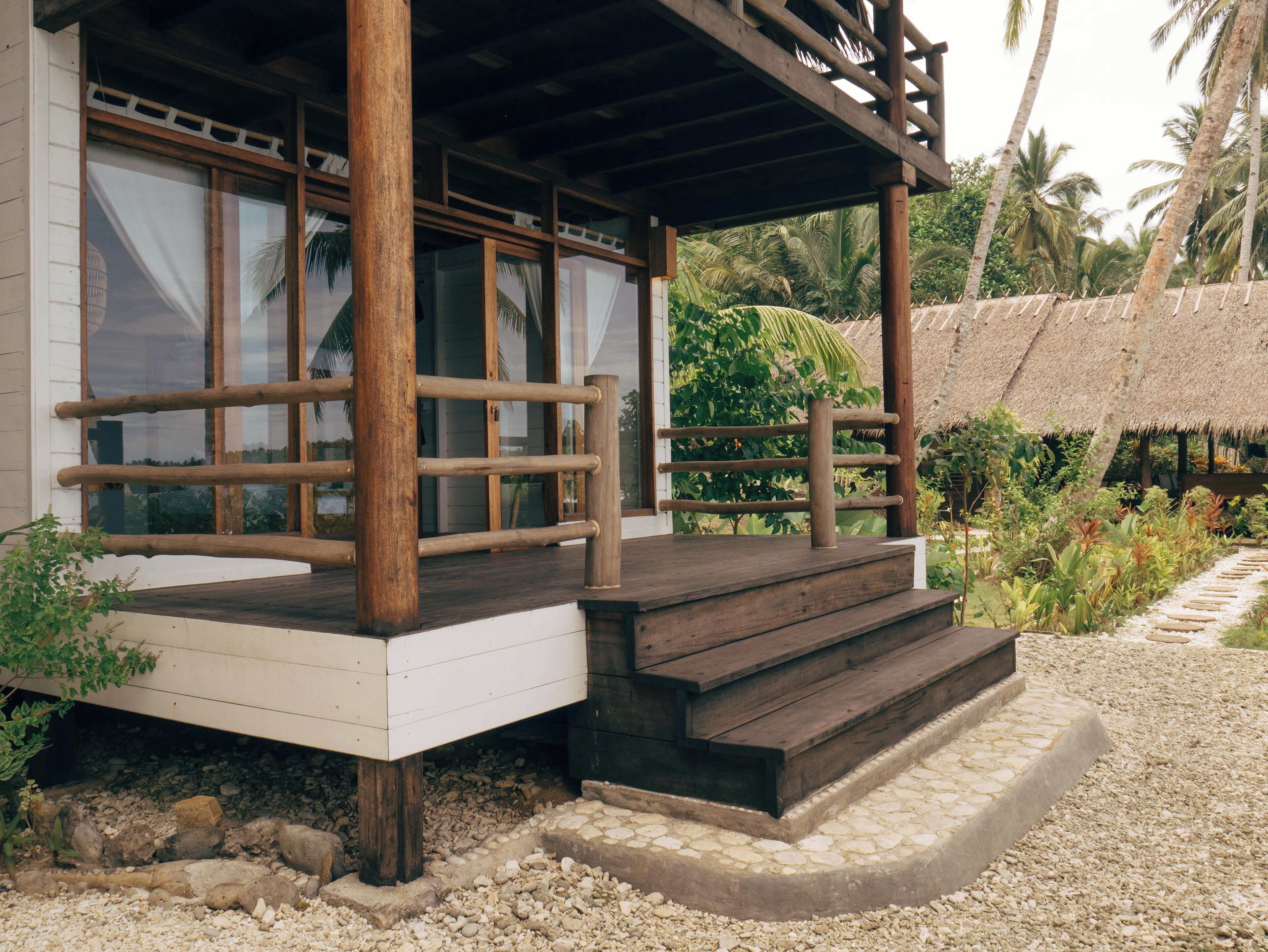 A wooden deck with steps leading to a covered area, surrounded by tropical vegetation and a thatched-roof structure.