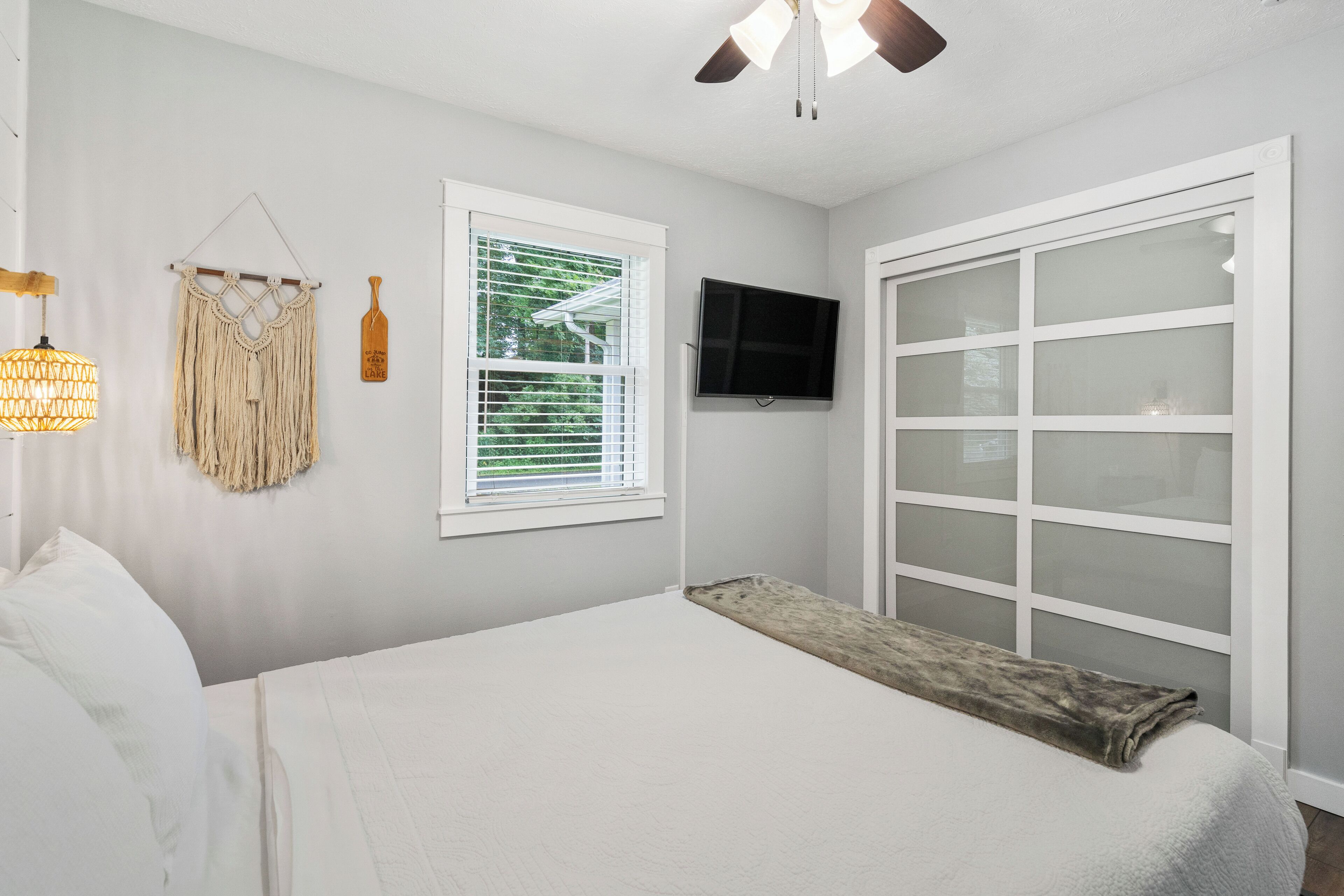 Une chambre à coucher avec un lit, une fenêtre avec des stores, un téléviseur fixé au mur et un ventilateur de plafond.