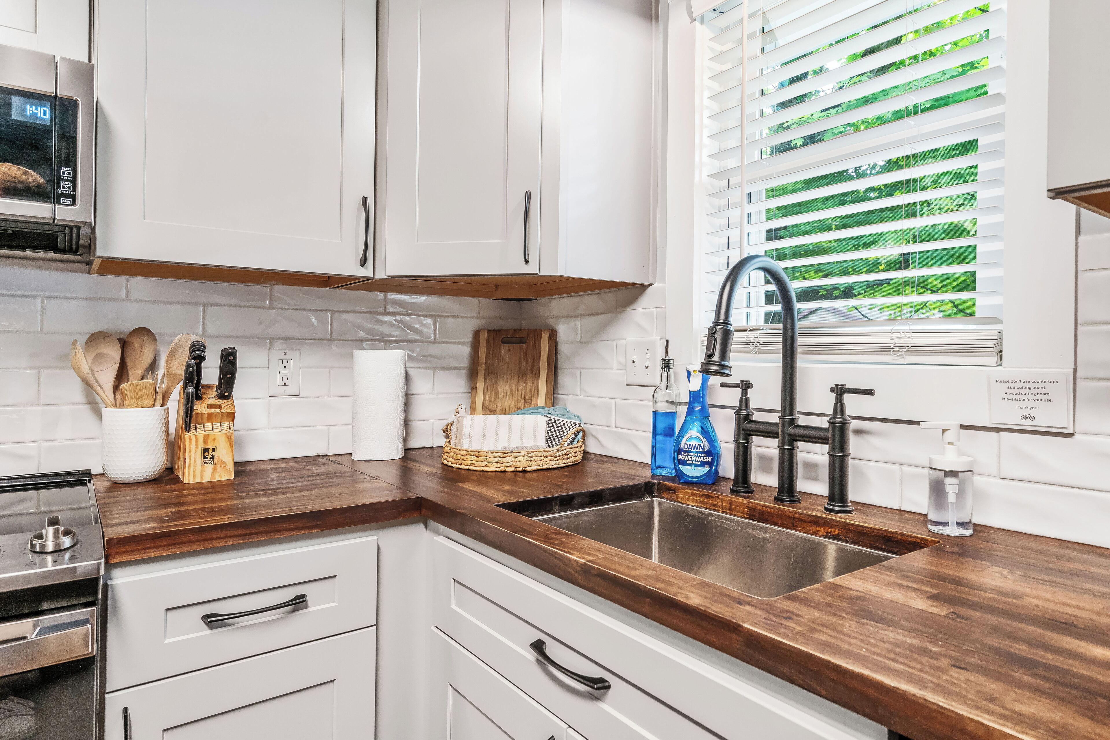 A modern kitchen with white cabinets, a wooden countertop, a stainless steel sink, and a window with blinds.