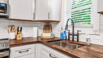 A modern kitchen with white cabinets, a wooden countertop, a stainless steel sink, and a window with blinds.