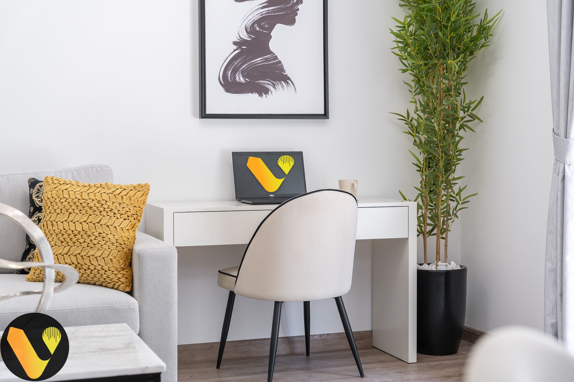 A modern office space with a white desk, a laptop, a framed artwork, a potted plant, and a sofa with a yellow cushion.