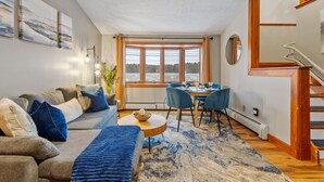 Un salon moderne avec un canapé, une table à manger et des chaises. Une grande fenêtre offre une vue sur des arbres et un lac. Un escalier est visible sur le côté droit.