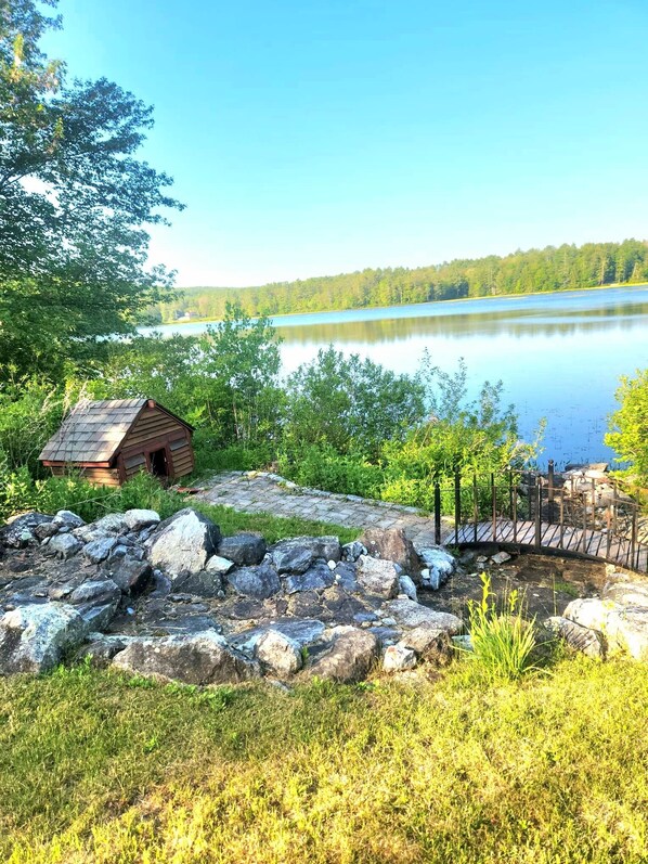 Una cabaña rústica junto a un lago, con una terraza de madera y una cerca de metal.