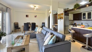 A modern living room with a grey sofa, a dining area with chairs, a kitchen with dark cabinets and a white countertop, and a large window with curtains. - Corfu SeaTy Apartment (Corfu)