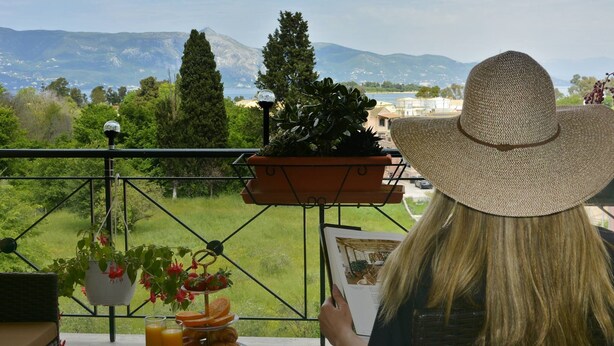 A balcony with a view of mountains, a potted plant, and a table with a drink and a book. - Corfu SeaTy Apartment (Corfu)