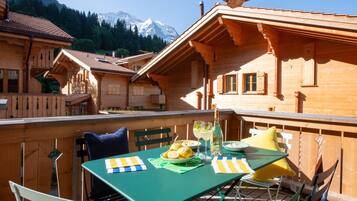 A wooden chalet with a balcony, a table with chairs, and a mountain view in the background.