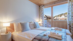 A double bed with white linens, a bedside table with a lamp, and a view of a town through a window.