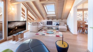 A cozy loft-style living room with a large TV, wooden beams, a sofa, and a dining area.