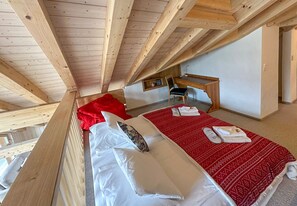 A bedroom with a sloped ceiling, wooden beams, a desk, a chair, and a bed with a red blanket and white pillows.