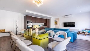 Un salon moderne avec une table à manger en verre, des chaises blanches, un canapé bleu et un coin cuisine doté de meubles en bois et d’un téléviseur.