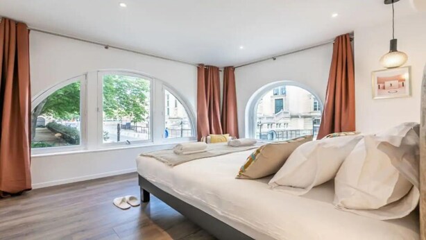 Une chambre d’hôtel moderne avec un grand lit, deux fenêtres avec des rideaux, une vue sur la ville et un tableau encadré au mur.
