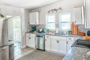 Una cocina moderna con armarios blancos, un refrigerador de acero inoxidable, un lavavajillas y una encimera de granito. Hay dos ventanas que ofrecen luz natural y vistas al exterior.
