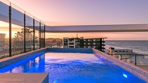 Ein Infinity-Pool auf dem Dach mit Blick auf das Meer und die Stadtlandschaft bei Sonnenuntergang.