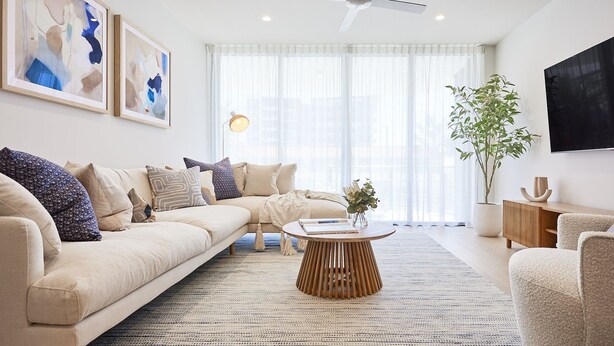 Ein modernes Wohnzimmer mit einem beigefarbenen Sofa, einem runden Couchtisch und einem großen Fenster mit leichten Vorhängen.
